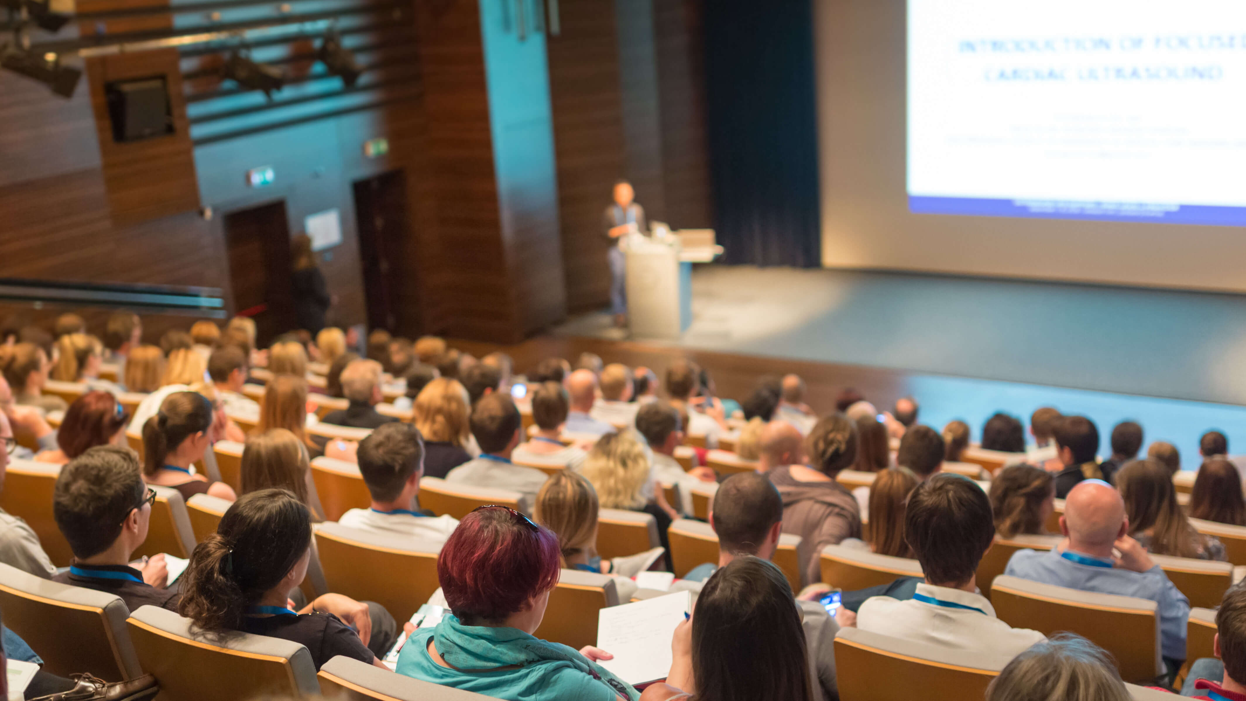 Audience seated in a lecture hall, watching a speaker at a podium with a presentation screen.
