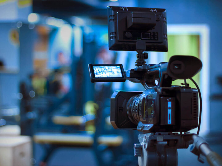 A professional video camera on a tripod in a blurred indoor setting.